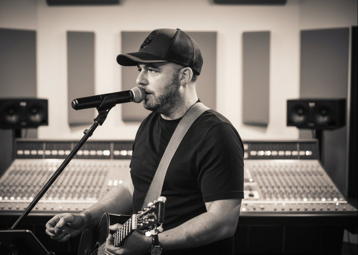 Man singing into a microphone with a guitar in a recording studio.