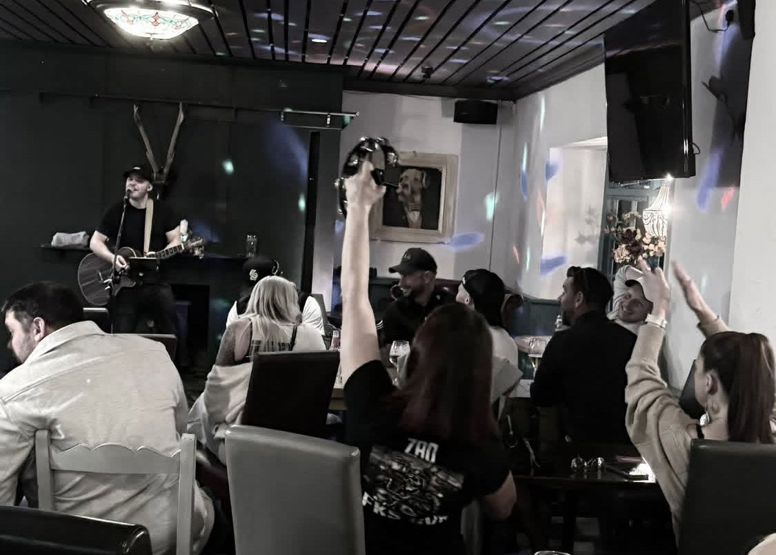 People in a bar with a guitarist on stage, raising their glasses.