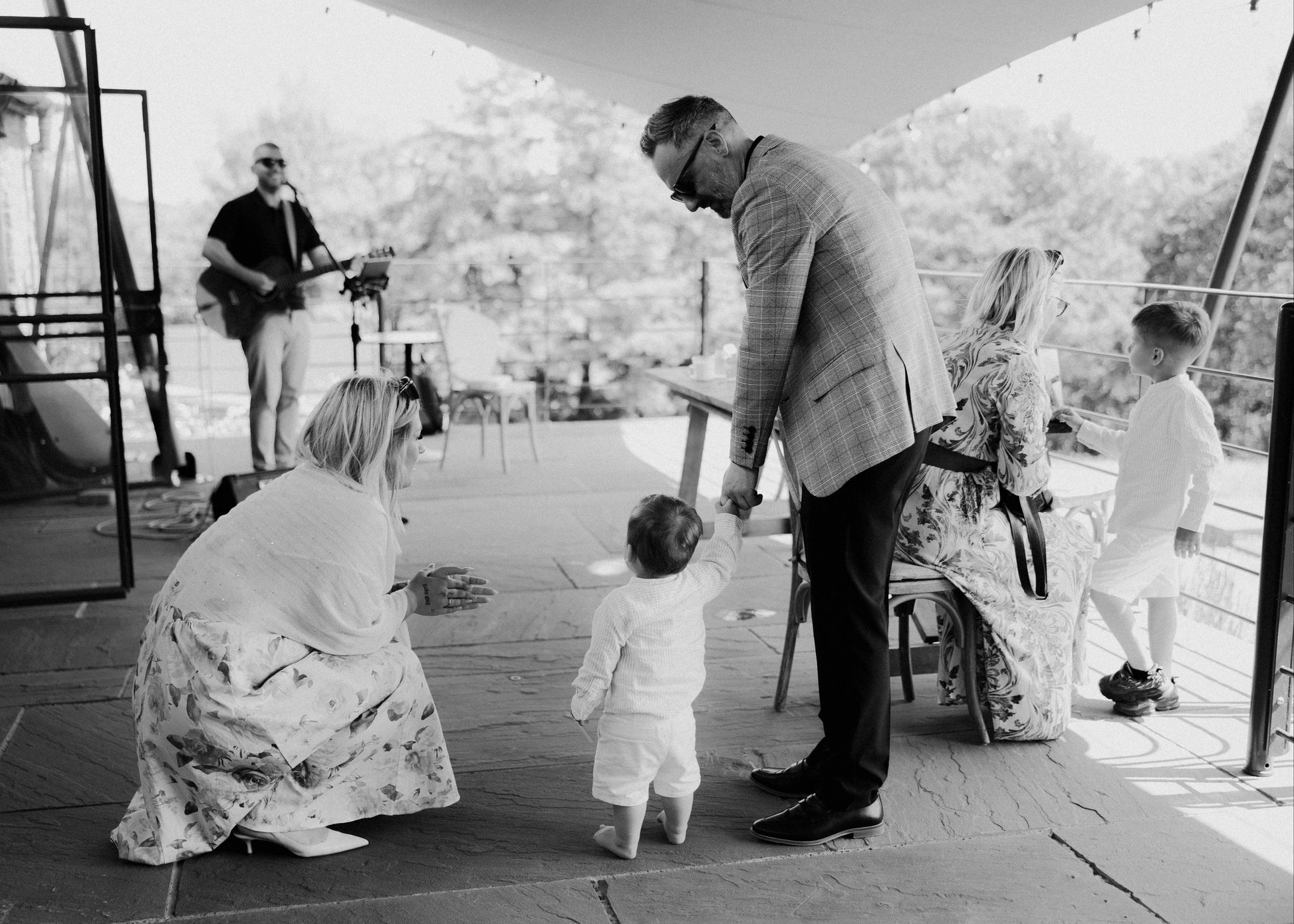Black and white photo of a family on a deck with a guitar player in the background